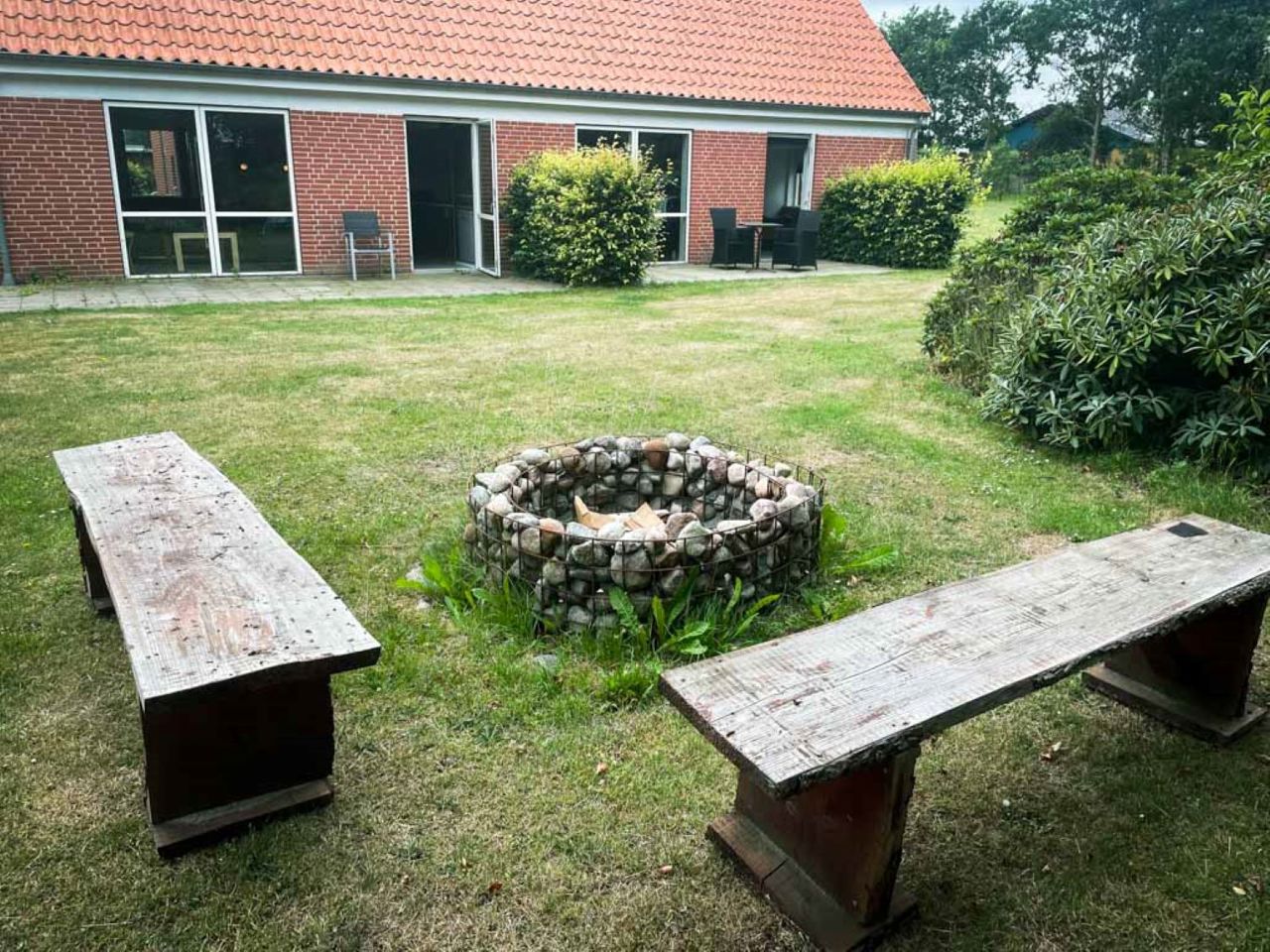 Rundt bålsted i haven med to bænke omkring og i baggrunden kan man se Lærkely, en rød bygning med hvide vinduer.