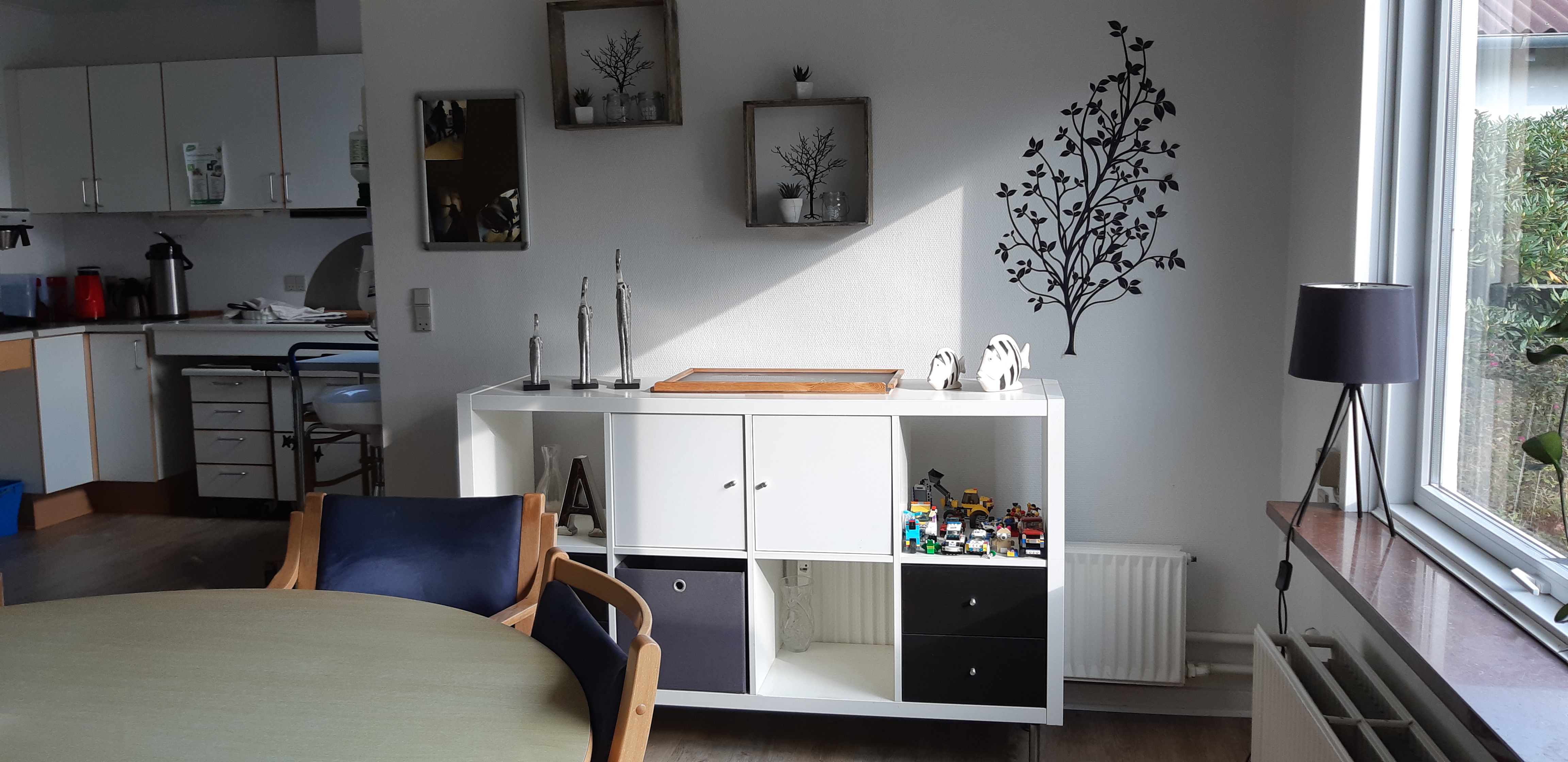 Fællesrummet hvor der er et køkken i baggrunden og et rundt spisebord med stole og en reol ved siden af.