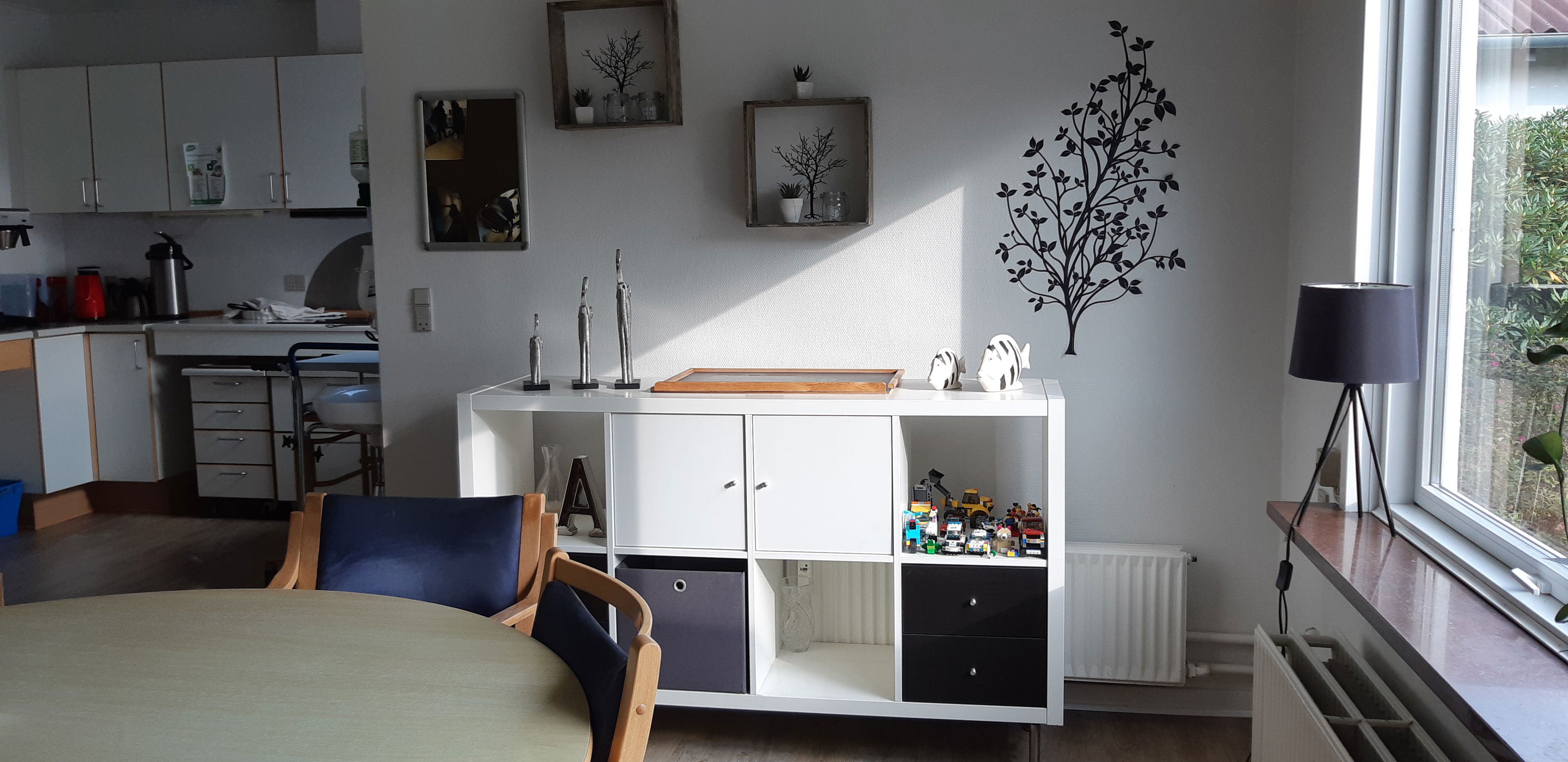 Fællesrummet hvor der er et køkken i baggrunden og et rundt spisebord med stole og en reol ved siden af.