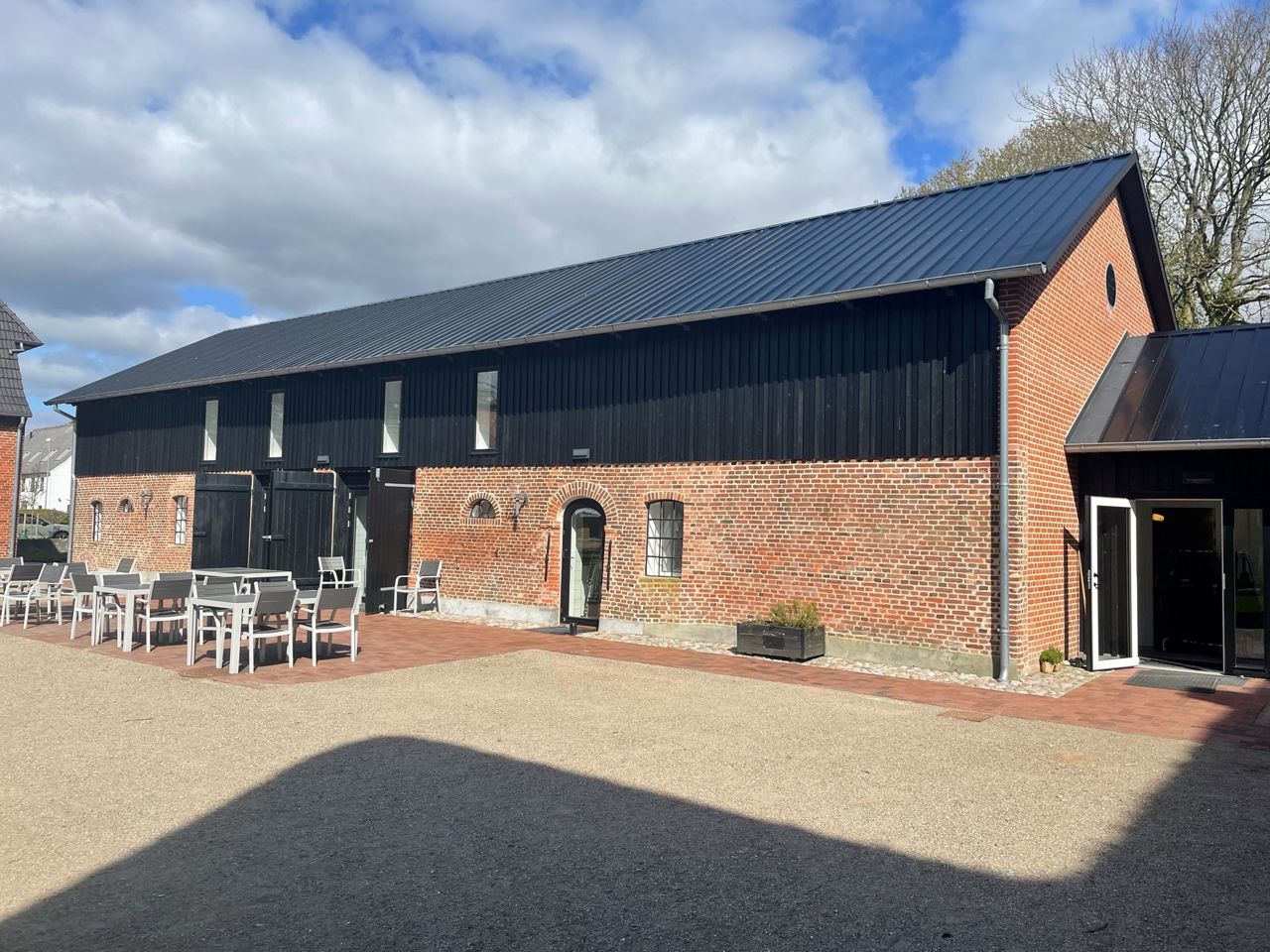 Smuk gårdsplads belagt med brosten i halvdelen og røde fliser i resten. Bagerst er der en bygning muret i røde mursten, og med sort tag. Foran bygningen står der flere sær havemøbler man kan sidde i.