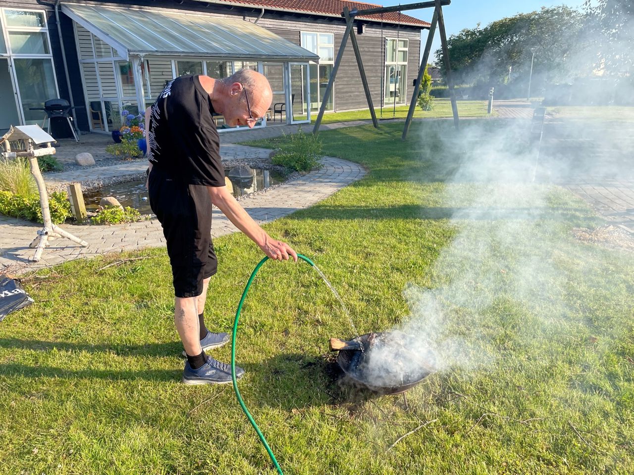 En borger står med en vandslange og hælder vand på et bål i et bålfad, rund om ham er der grønt græs og i baggrunden ses en bygning i sort træ.