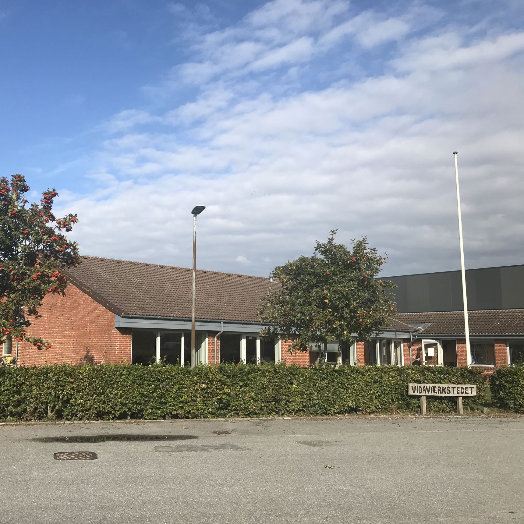 Indgangen til Vidåværkstedet. Bygningen er af røde mursten, og taget er af sort tegl. Til venstre i billedet står et stort træ med blade og orange bær. Foran hele bygningen er der en grøn hæk. Forrest i billedet er der asfalt parkeringsplads.