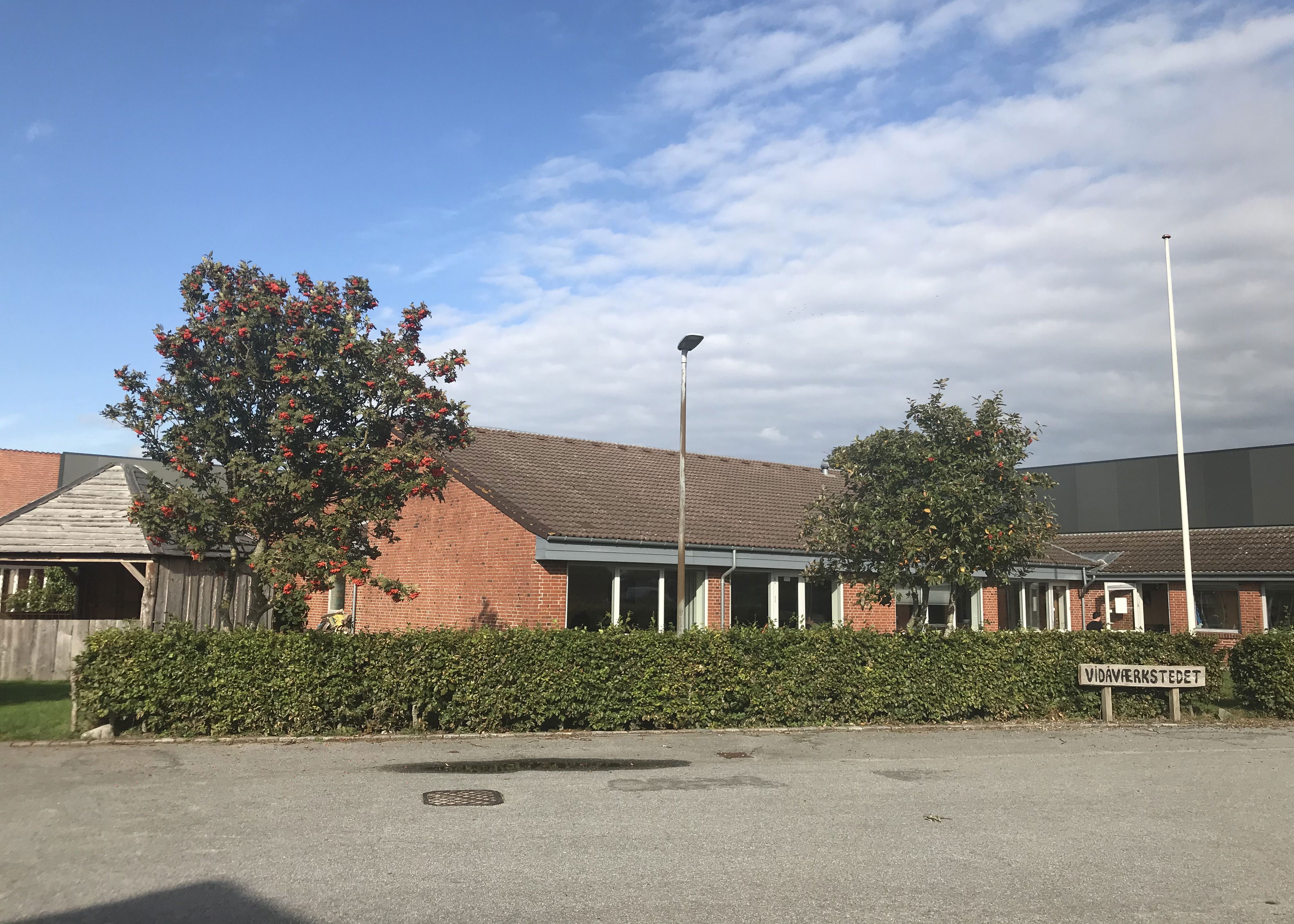 Indgangen til Vidåværkstedet. Bygningen er af røde mursten, og taget er af sort tegl. Til venstre i billedet står et stort træ med blade og orange bær. Foran hele bygningen er der en grøn hæk. Forrest i billedet er der asfalt parkeringsplads.