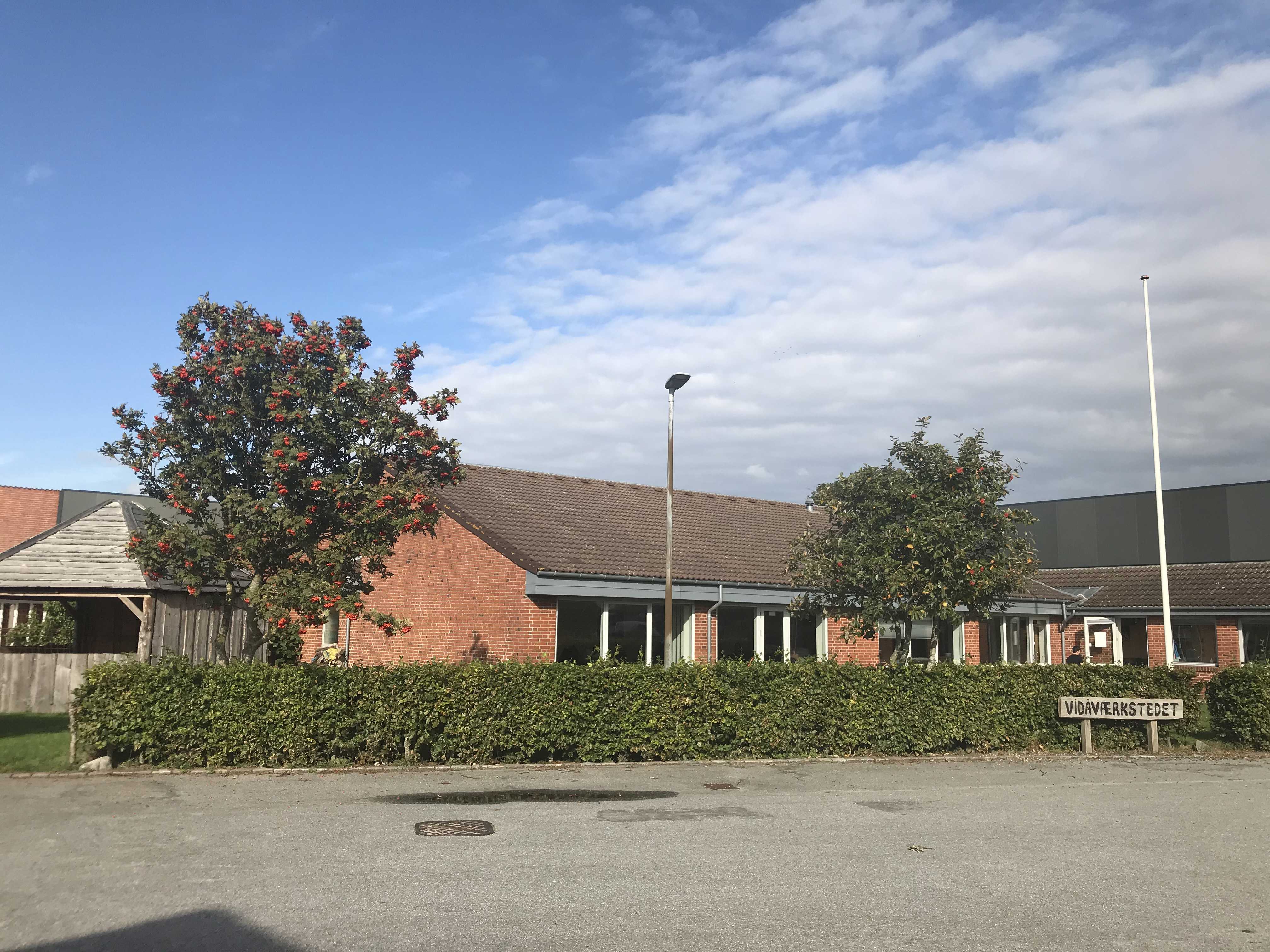 Indgangen til Vidåværkstedet. Bygningen er af røde mursten, og taget er af sort tegl. Til venstre i billedet står et stort træ med blade og orange bær. Foran hele bygningen er der en grøn hæk. Forrest i billedet er der asfalt parkeringsplads.
