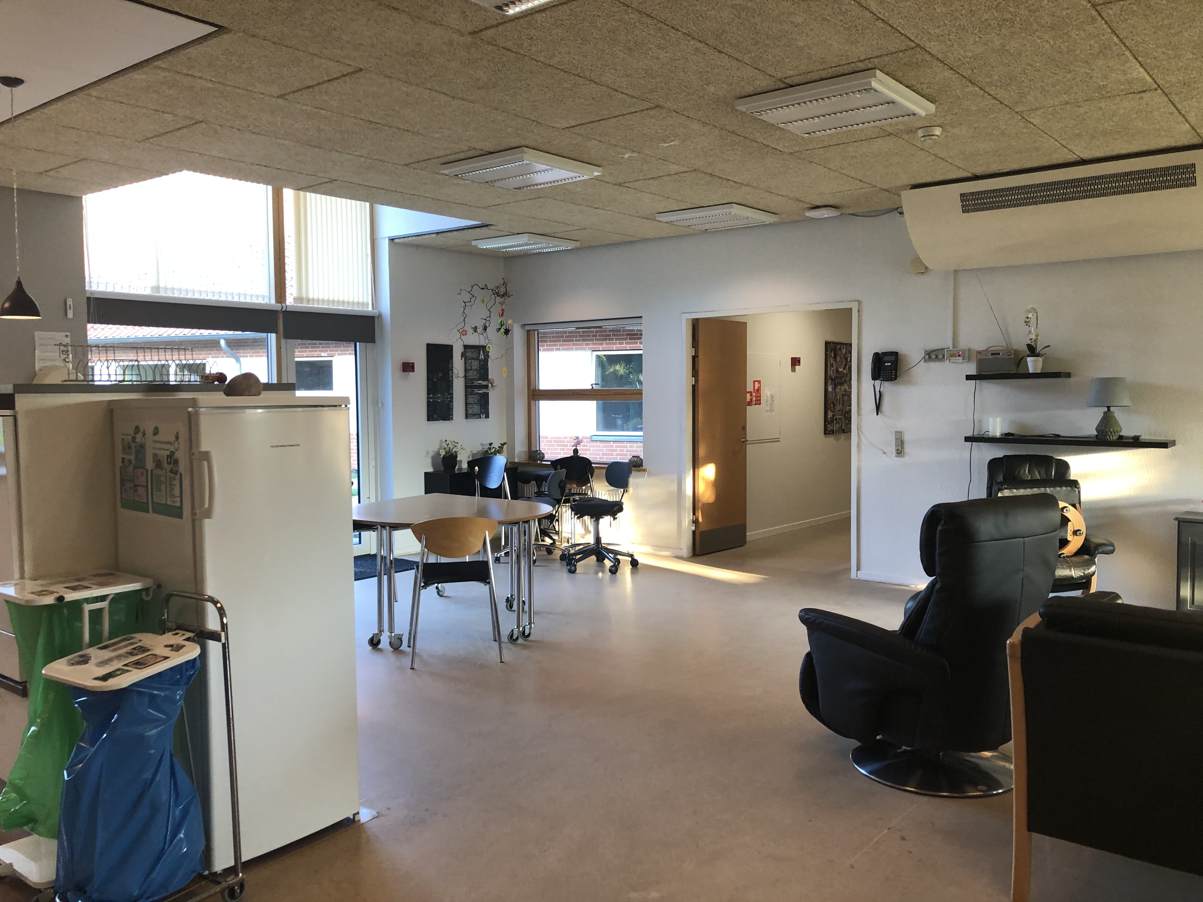 I opholdsrummet der står et lille bord  med nogle stole omkring. Derudover er der en læderlænestol og man kan se et køleskab.