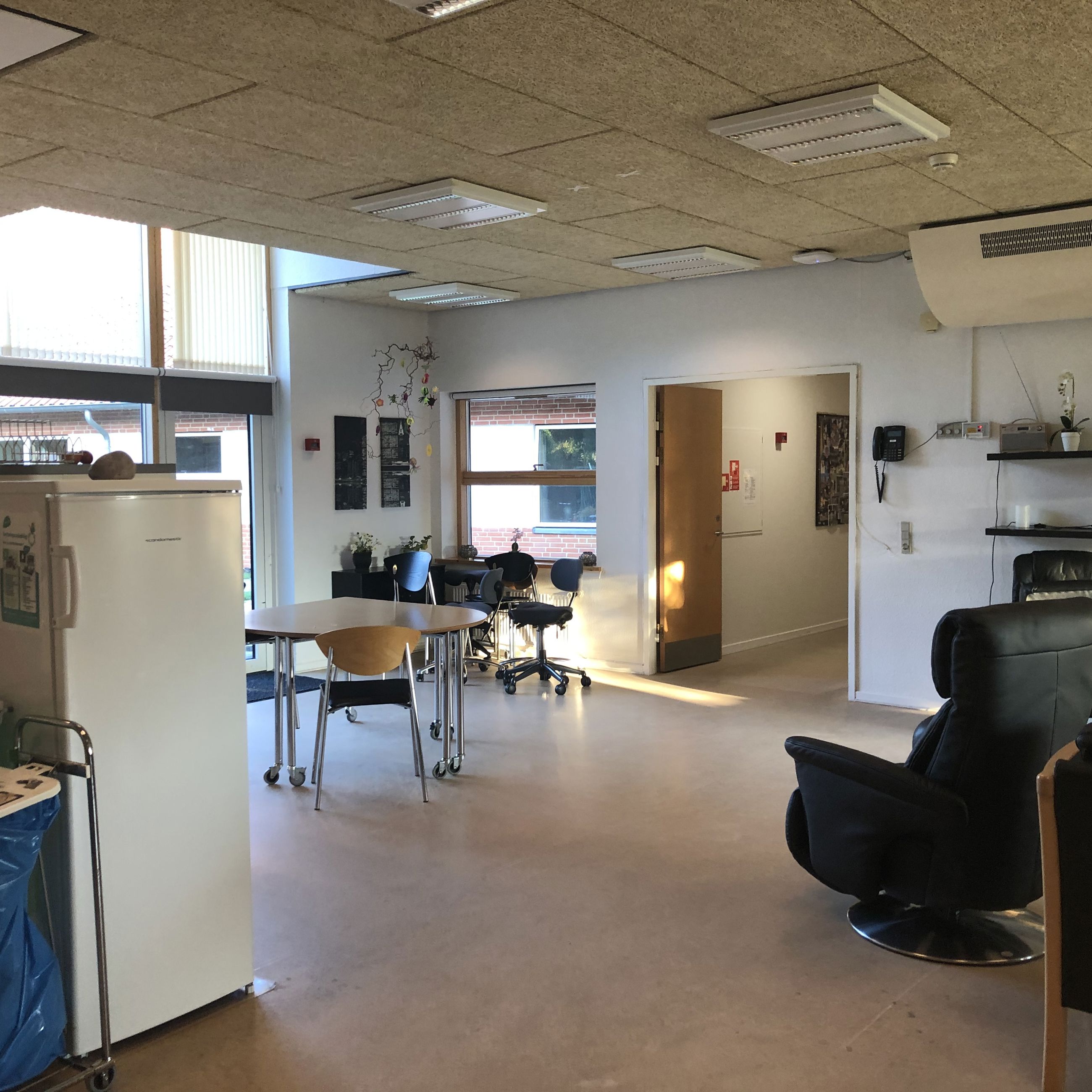 I opholdsrummet der står et lille bord med nogle stole omkring. Derudover er der en læderlænestol og man kan se et køleskab.