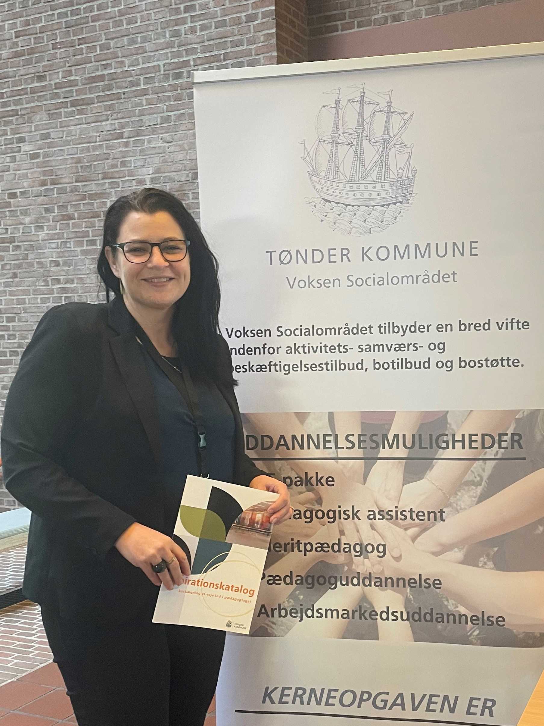 Der står en kvinde på billedet, hun står foran en stander der bliver brugt til jobmesse. Kvinden er klædt i sort tøj. På standeren står der forklaret hvad Tønder Kommune kan tilbyde dem der bliver uddannet pædagog.