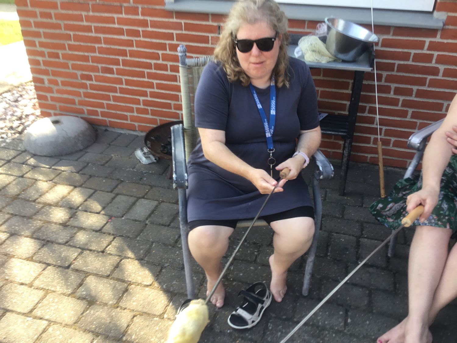 To borgere der sidder udenfor og laver snobrød, de sidder på havestole på en terrasse af fliser, det ser hyggeligt ud.