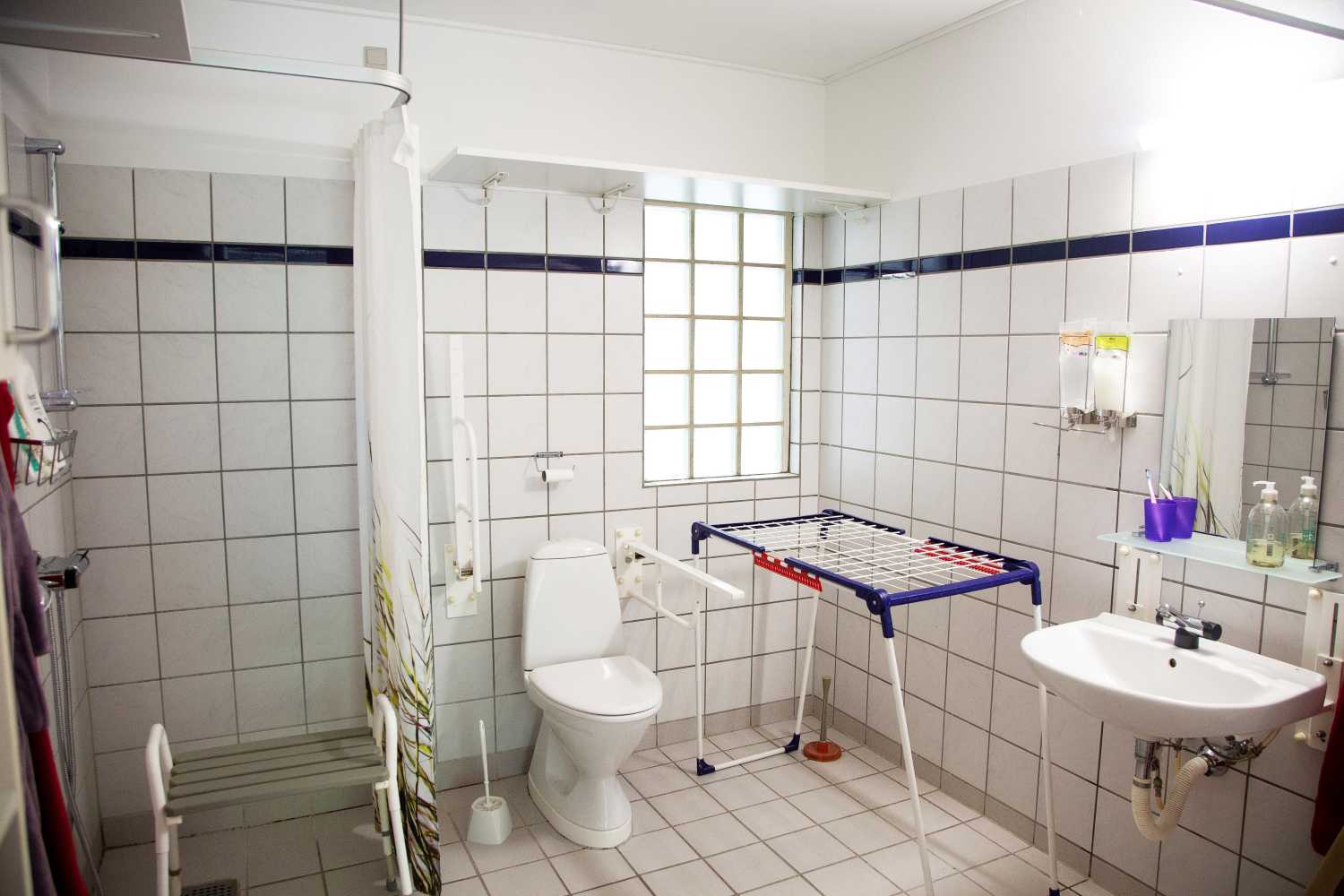 Badeværelse med toilet og vask og et brusehjørne med forhæng og i hjørnet står der et tøjstativ.