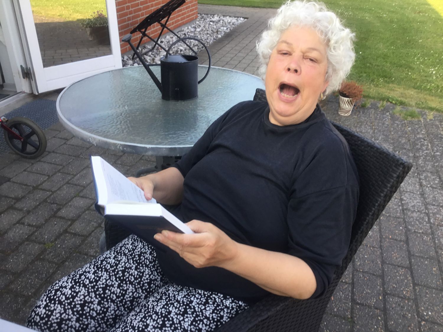 En borger sidder ved et havebrod i en havestol udenfor på fliseterrassen og synger, hun ser glad ud.
