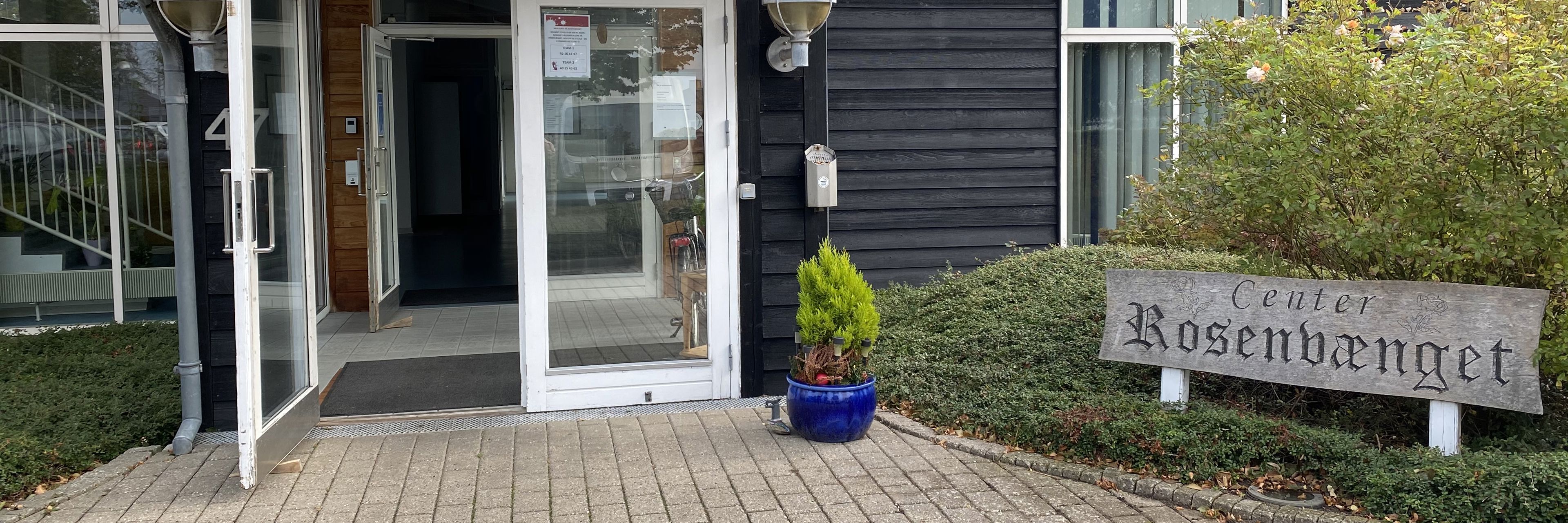Hovedindgang til Rosenvænget Botilbud med dobbelt glasdør. Bygningen er af sort træ, og der er rødt tegl tag på. Til højre for døren står der en krukke med en grøn plante i. Bedet der er lige ved siden af er grønt, og der står et træskilt hvor der står Center Rosenvænget