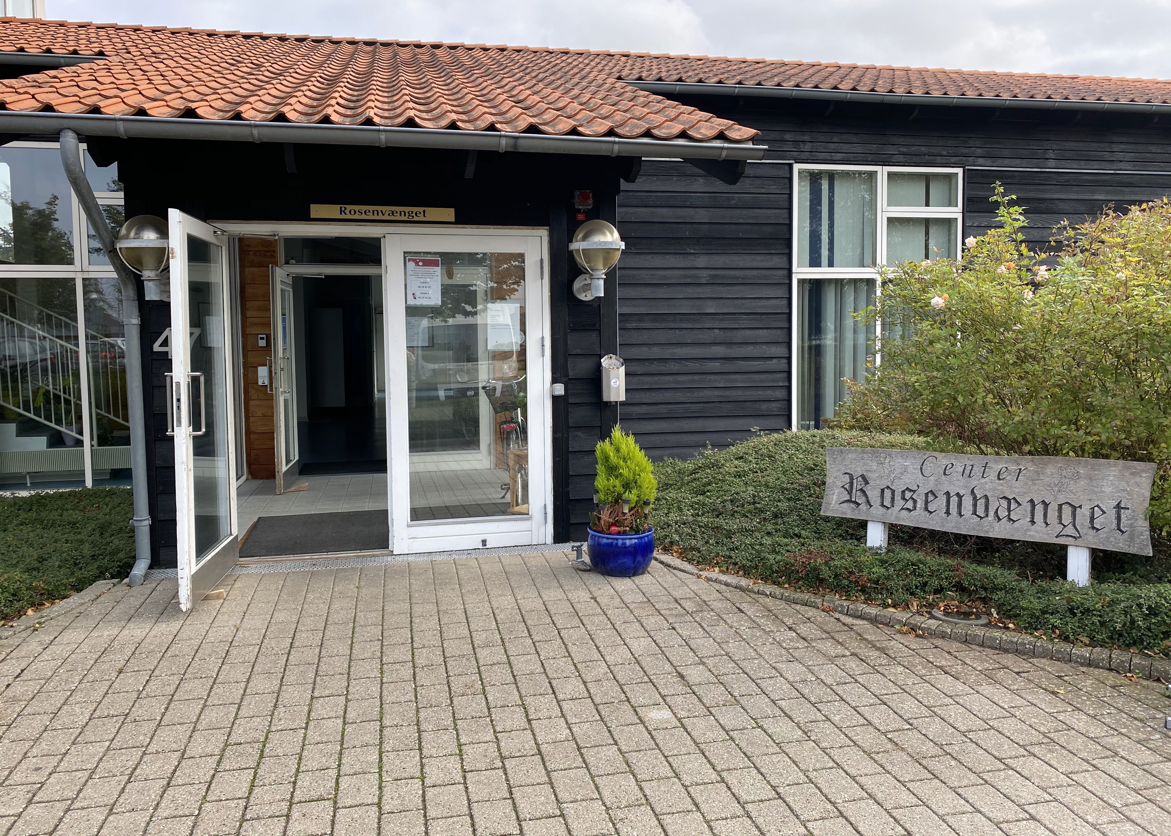 Hovedindgang til Rosenvænget Botilbud med dobbelt glasdør. Bygningen er af sort træ, og der er rødt tegl tag på. Til højre for døren står der en krukke med en grøn plante i. Bedet der er lige ved siden af er grønt, og der står et træskilt hvor der står Center Rosenvænget