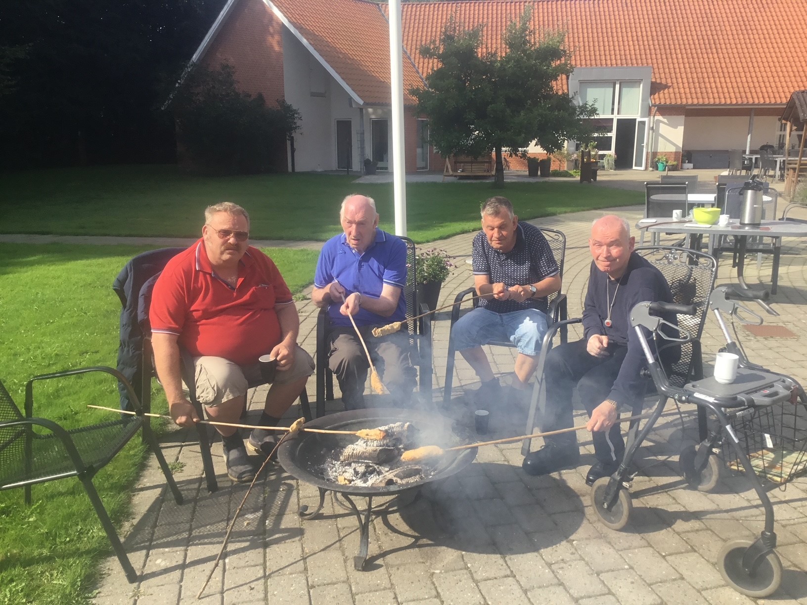 Fire borgere sidder ved et bålsted med hver deres snobrodspind med snobrød på, i baggrunden kan man se en stor bygning
