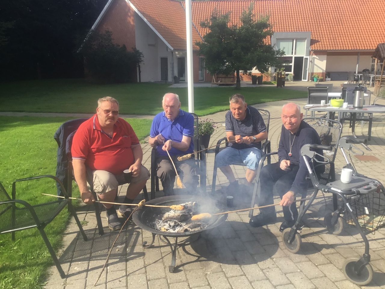 Fire borgere sidder ved et bålsted med hver deres snobrodspind med snobrød på, i baggrunden kan man se en stor bygning