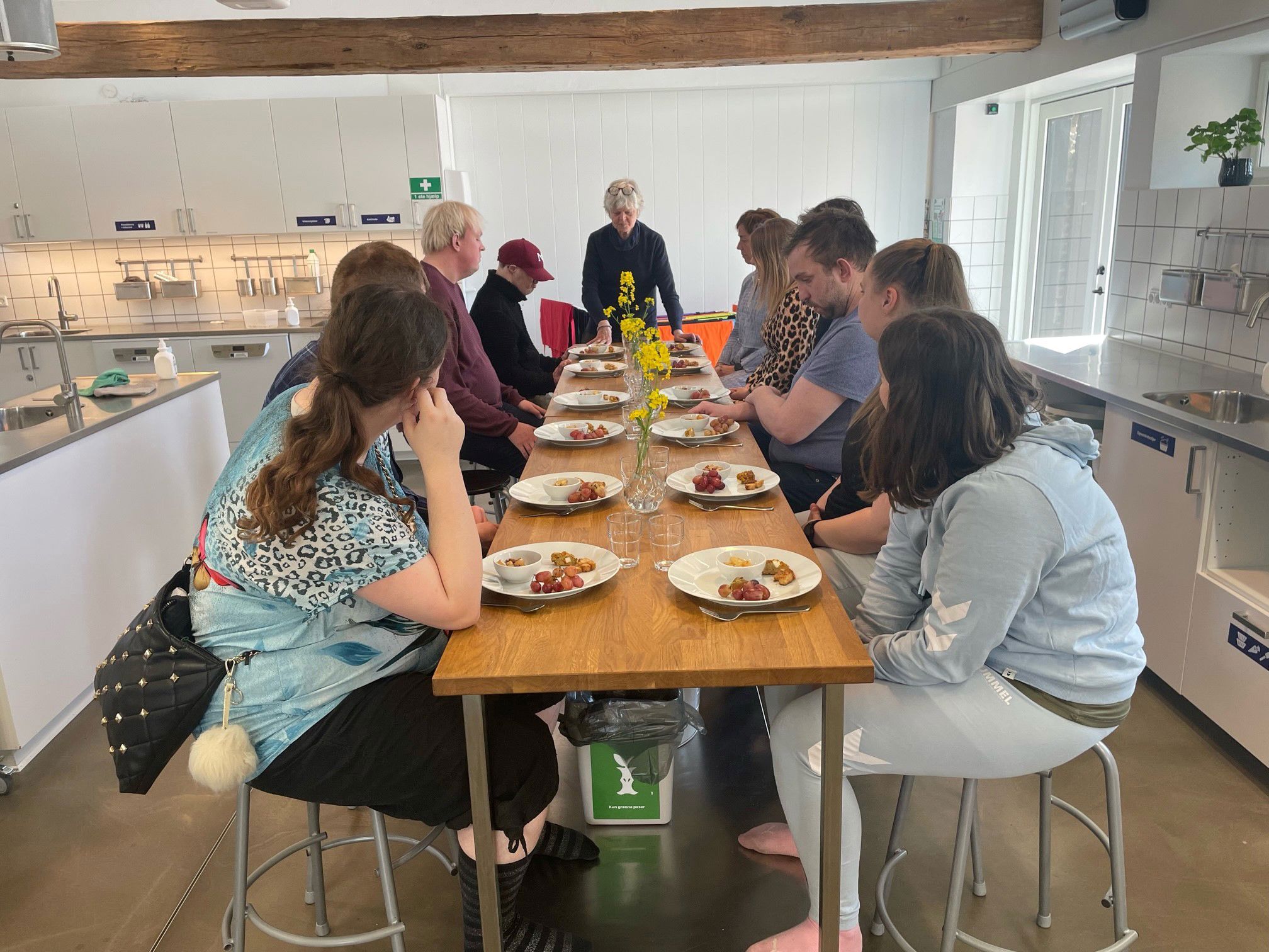 Ti personer sidder ved et langt bord, hvor bordpladen er af træ. De ti personer sidder og hører et foredrag. I den ende af bordet står en kvinde og taler.