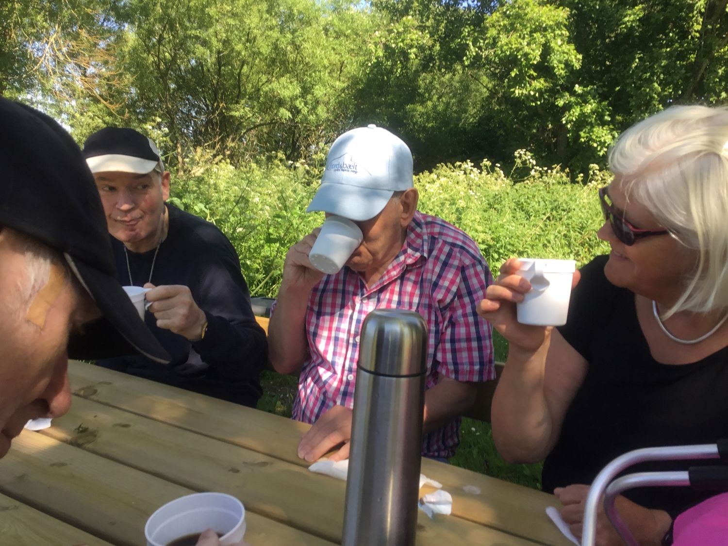 Flere borgere sidder ved et bord ude i solskind og får en kop kaffe, de ser glade ud, i baggrunden er der grønne træer og buske