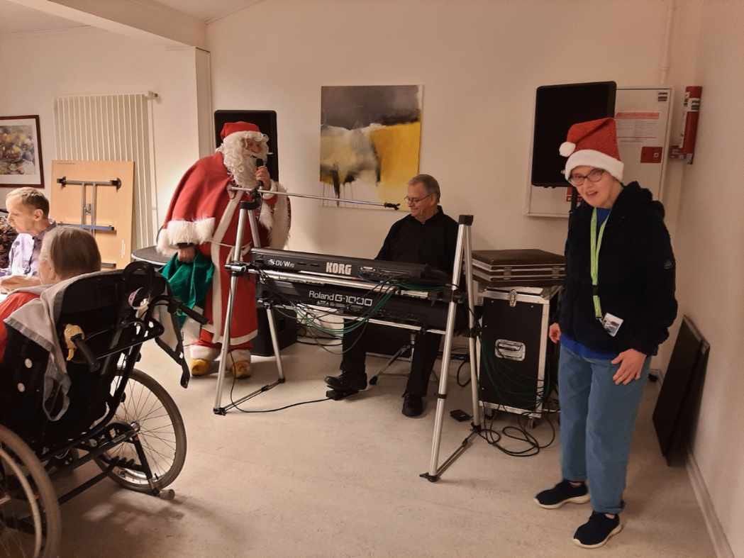 Julemanden står og giver et nummer, der sidder en mand ved et keyboard og spiller. I forgrunden står der en borger og danser.