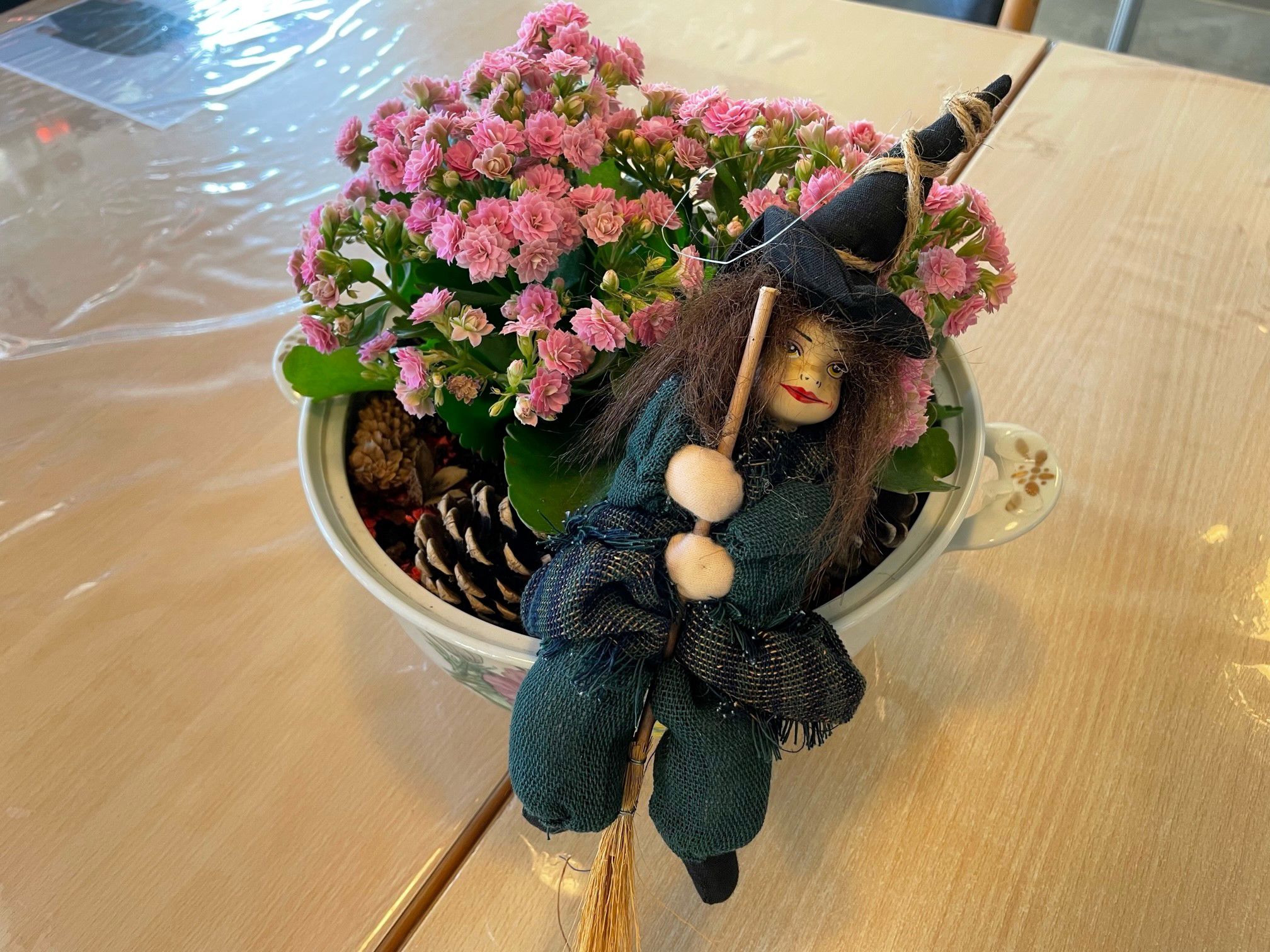 En blomster dekoration med lyserøde blomster i, grankogler og en pynteheks den står på et træbord med en gennemsigtig voksdug på