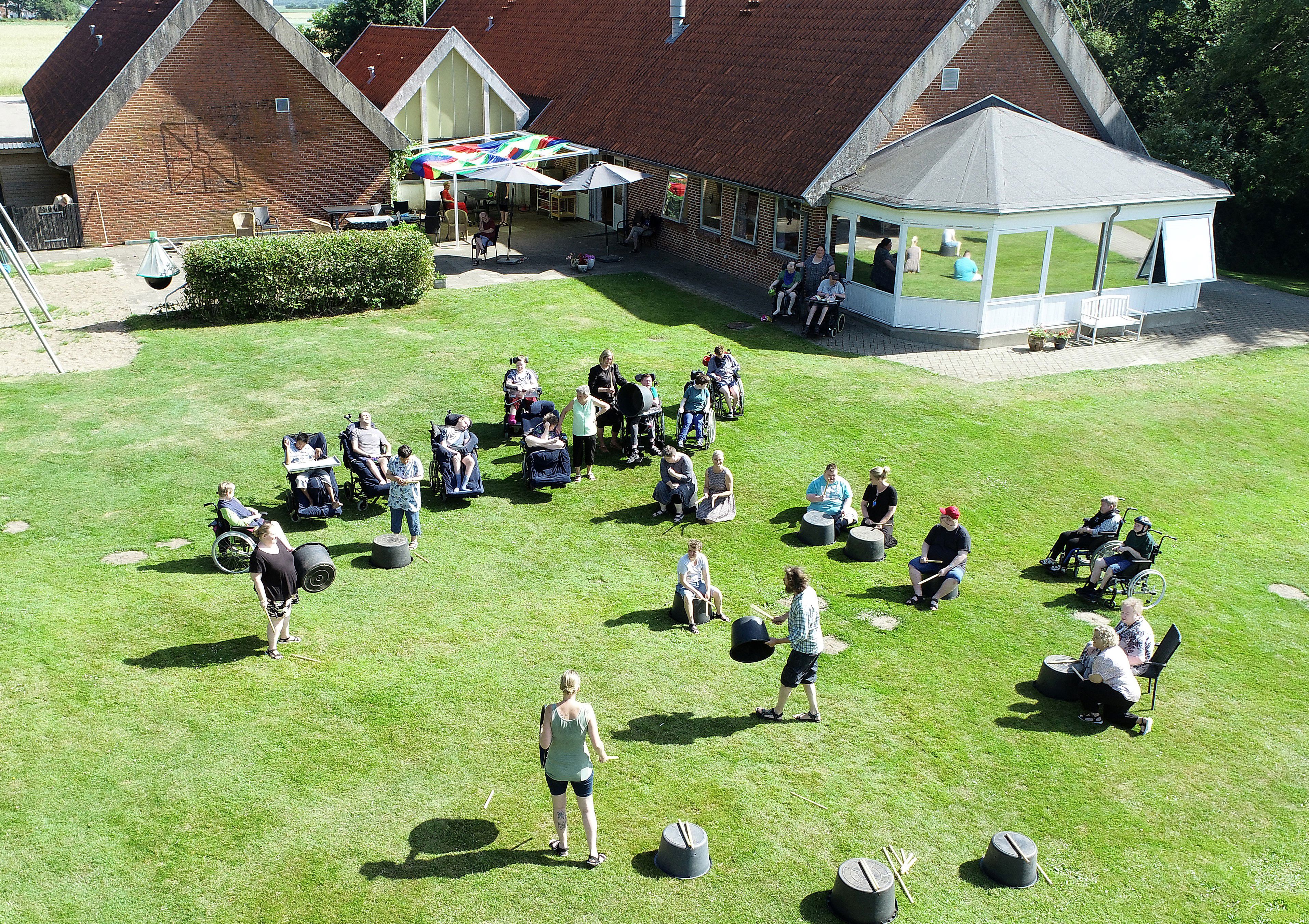 Foto taget oppefra af borgere der står spredt på en græsplæne med nogle baljer, nogle står på baljerne med trommestikker, bagved ses bygningen med rode mursten og et faverigt soldækken over terrassen.