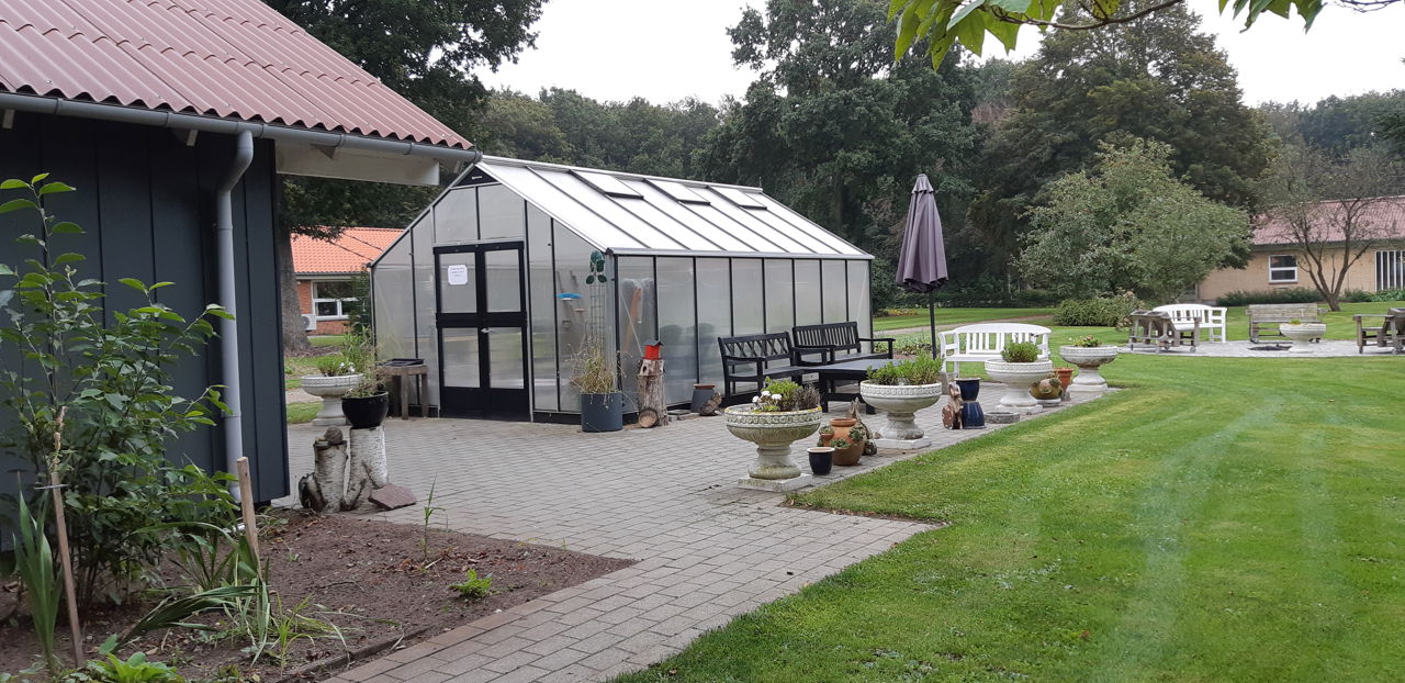 Der er et stort drivhus som ligger på en flisebelagt grund og rundt om er der en stor græsplæne, der er bænke hvor man kan sidde.