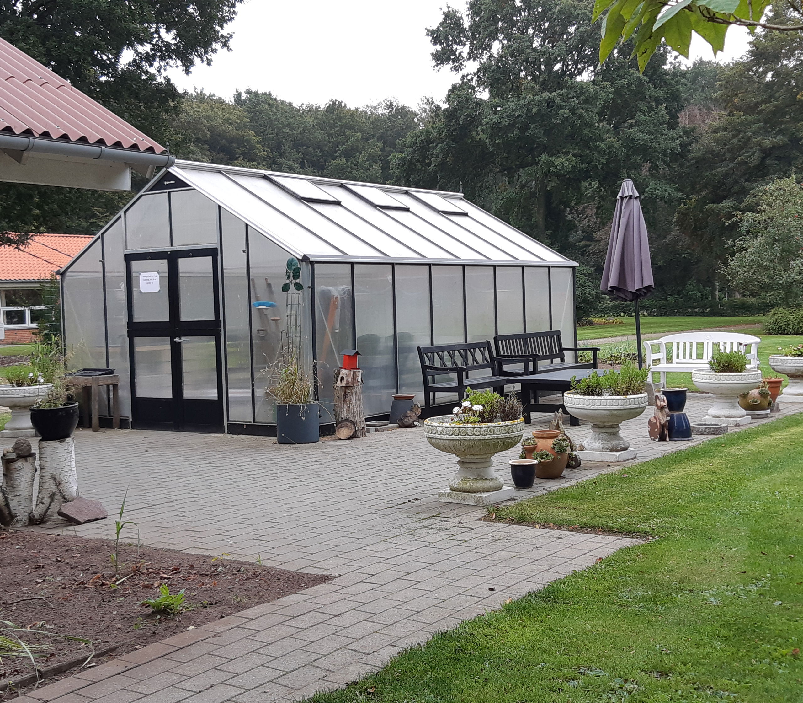 Der er et stort drivhus som ligger på en flisebelagt grund og rundt om er der en stor græsplæne, der er bænke hvor man kan sidde.