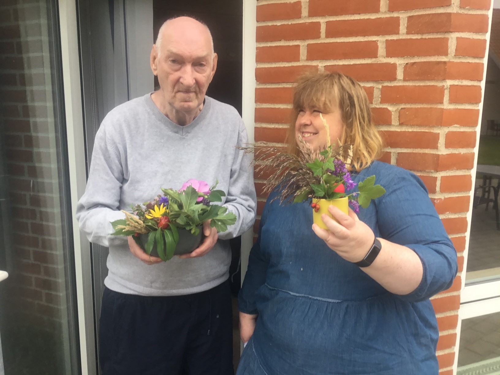 To borgere viser flotte blomster frem, de står foran en glasdør i en en bygning med røde mursten.