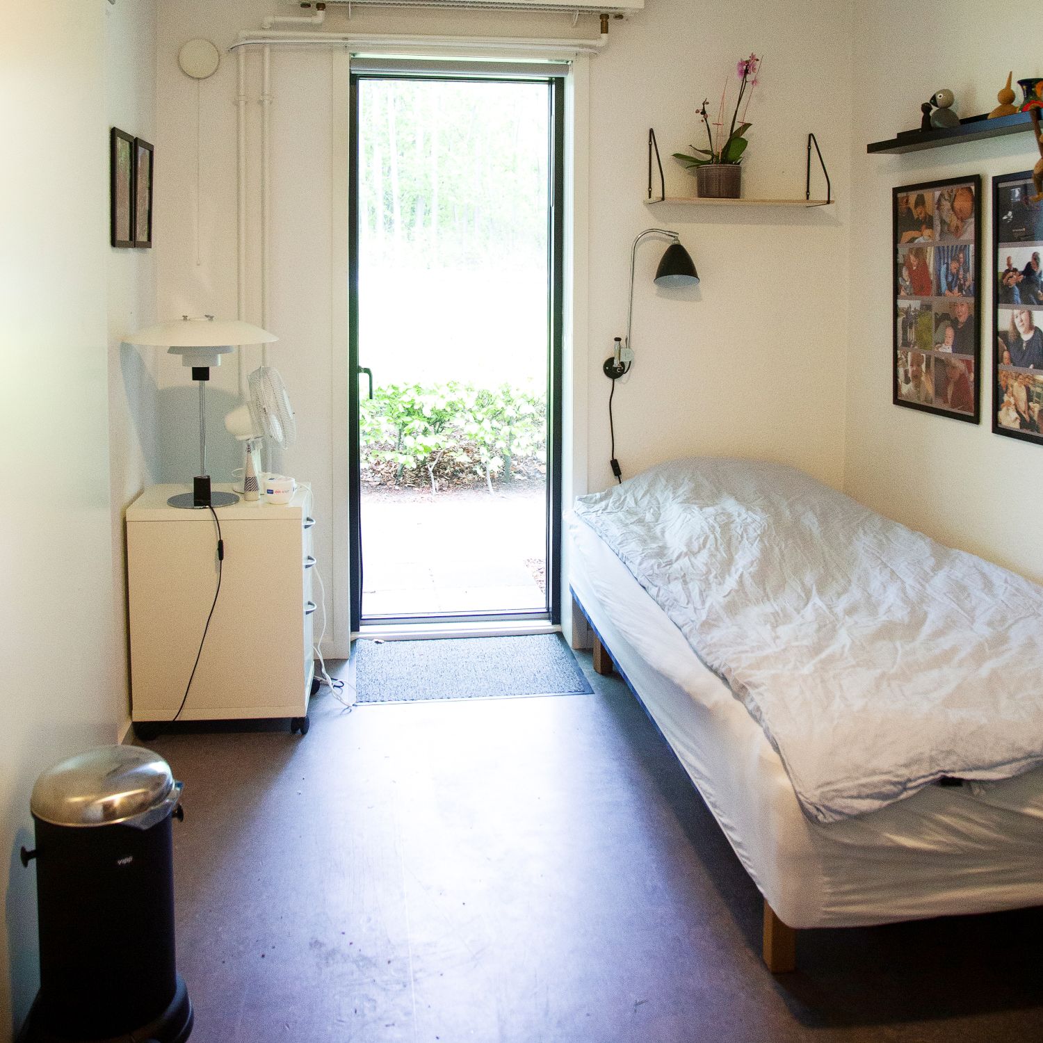 soveværelse i lejlighed hvide vægge en seng med hvidt sengetøj en havedør for enden af rummet og overfor en kommode med en lampe på