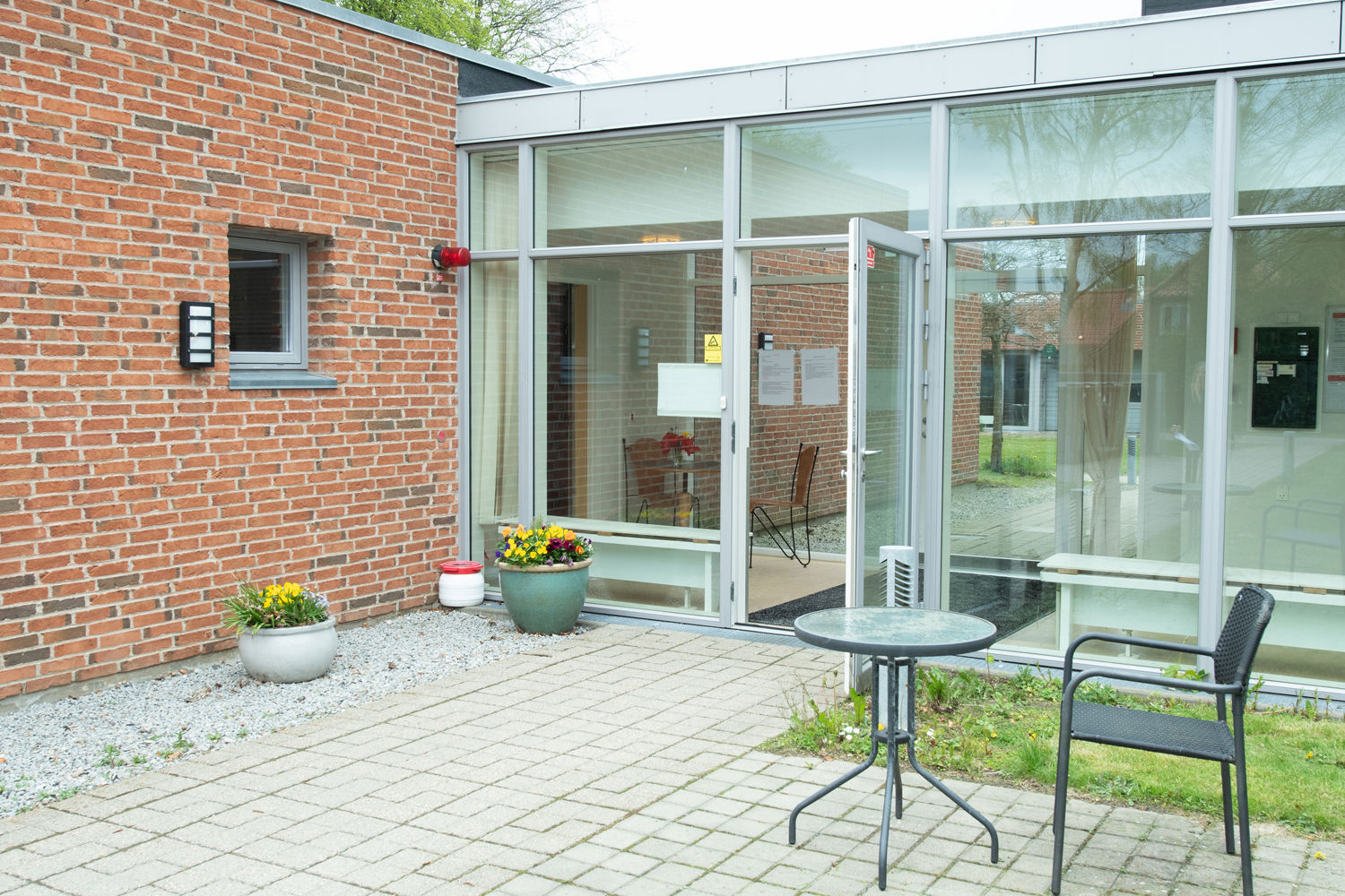 Ved indgangen står der et cafébord og en sort havestol. Der står to krukker med blomster.